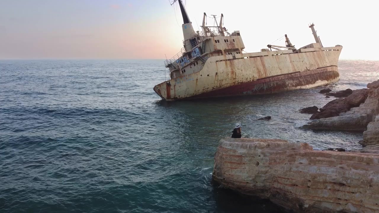 Una nave da carico abbandonata si trova incagliata su rocce costiere con onde delicate che lambiscono la riva sotto un cielo velato.