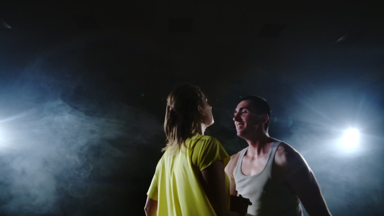 la cámara está zoomando y disparando desde el plano completo hasta el medio uno en mocking slow una joven con un vestido amarillo y un hombre con una camisa blanca bailando en el escenario bajo los reflectores y el humo