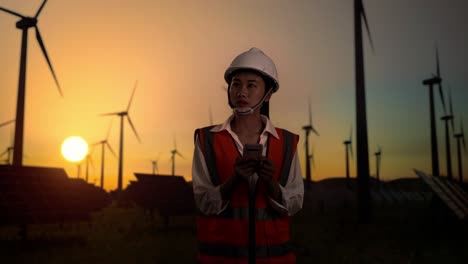 asian female engineer in a helmet standing in front of wind turbines rotating at sunset, using smartphone and looking around