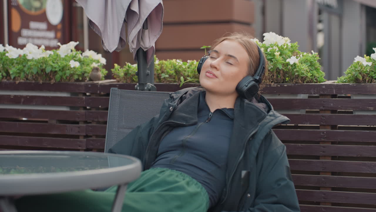 Ung kvinde lytter udendørs med hovedtelefoner, afslappet udtryk, hyggelig jakke, caféterrasse, træbænk, potteplanter, urban gade baggrund, lukkede øjne, subtilt smil, nyder ambient musik