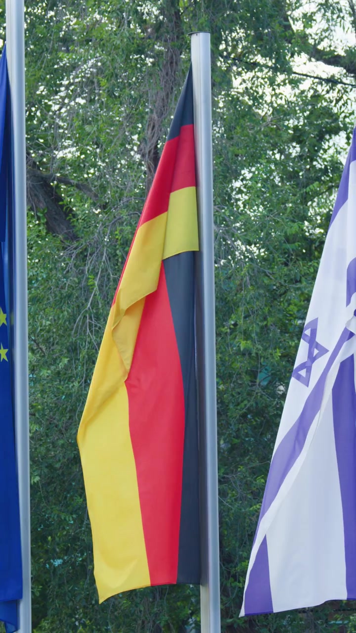 German, Israeli, and European Union flags waving in wind, outdoor setting, natural daylight, static shot