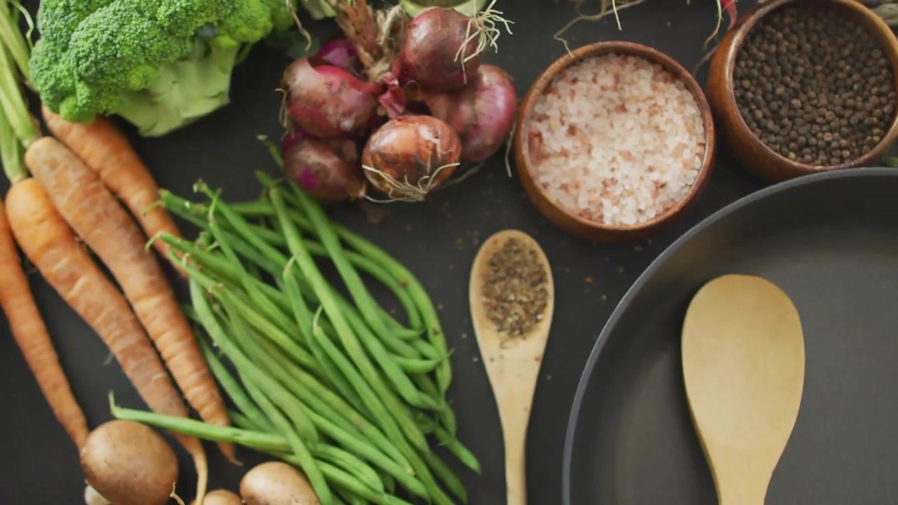 Composizione flat lay che si dispiega su un piano di lavoro scuro, con bagliori che evidenziano verdure e utensili per la preparazione della cucina.