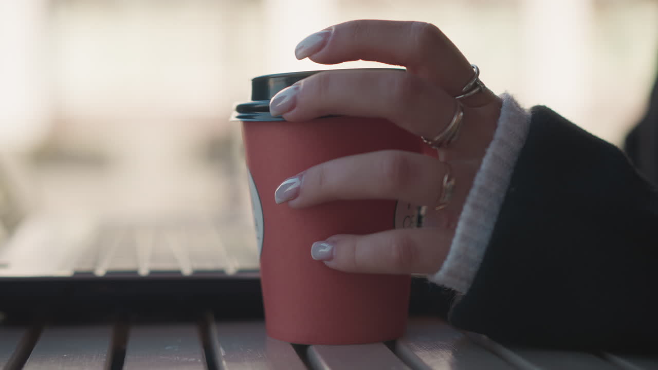 primer plano de la mano de la mujer sosteniendo una taza de café roja frotando suavemente la parte superior con uñas manicuradas mientras la computadora portátil se sienta en la parte delantera de una mesa al aire libre, fondo borroso suave