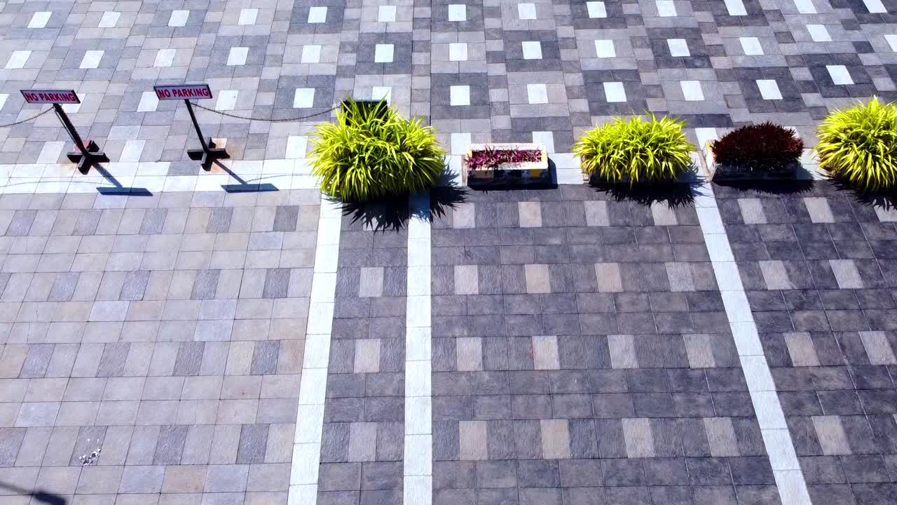 Aerial top-down view of a modern paved courtyard featuring geometric patterns, vibrant green shrubs in planters, and "No Parking" signs under bright sunlight