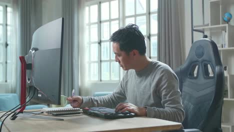 asian man in long sleeved t-shirt looking at desktop computer screen and writing in notebook on a table while working at home.