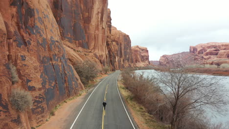 Vista-Aérea-De-Un-Hombre-Solitario-Corriendo-En-Una-Carretera-Desierta-Por-El-Río-Colorado-Bajo-Altos-Acantilados-De-Arenisca-Empinados-Del-Desierto-De-Utah,-Ee.uu.,-Seguimiento-De-Disparos-De-Drones