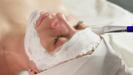 high angle view of gloved aesthetician carefully applying thick white mask with silicone brush along contours and corners of guest face on massage bed during relaxing spa facial treatment session