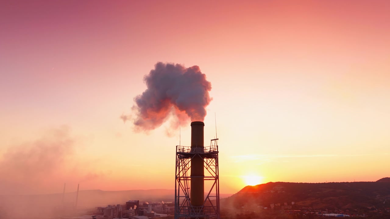 Struttura di camino a traliccio metallico con fumo al tramonto. Emissioni rilasciate in un cielo viola e giallo da un'alta condotta d'acciaio in un impianto industriale.