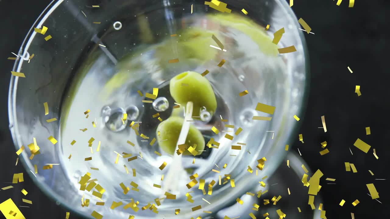Hand entering upper right stirring glass, forming vortex bubbles, olive dropping, confetti drifting