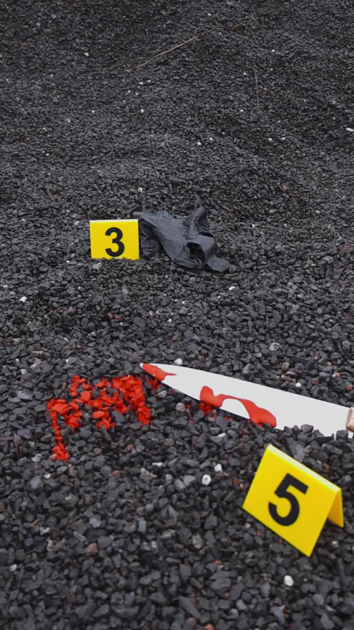 Crime scene police markers next to evidence on gravel ground outdoors in a vertical video. Bloody knife, black nylon glove is visible in the video that spins around them. Shallow depth of field