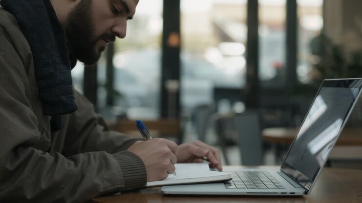 Glancing at laptop, student in jacket taking notes in notebook with blue pen at cafe