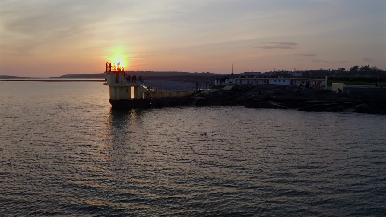 Ripresa aerea di presentazione di un molo e una torre mentre la luce svanisce e sfumature dorate si diffondono sul mare calmo, con persone che nuotano e stanno in piedi lungo la ringhiera