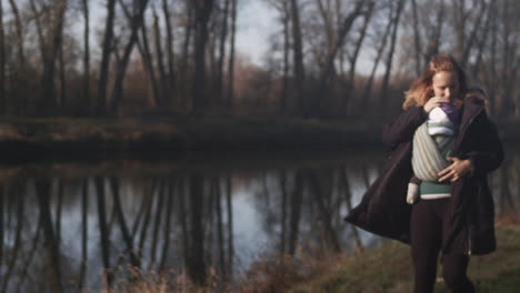 Mother-enters-frame-walking-with-baby-in-sling-beside-peaceful-lake