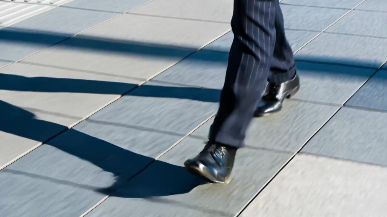 Uomo che fa un passo con il piede destro, camminando su un sentiero con striscia tattile, con pantaloni da completo e scarpe nere, per andare al lavoro.