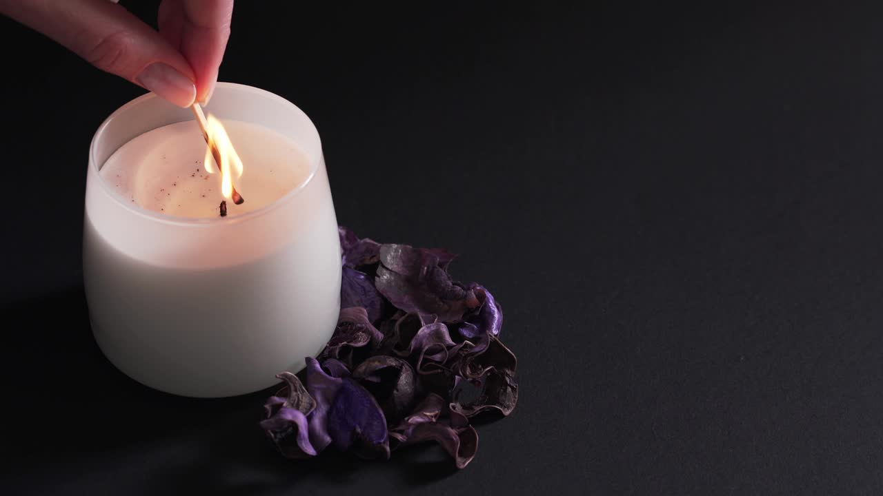 Aromaterapi konsepti - siyah arka plan üzerinde izole edilmiş beyaz bir mumu yakan bir kadın eli. Spa'da rahatlama ve meditasyon için kuru kokulu çiçekler 4K.