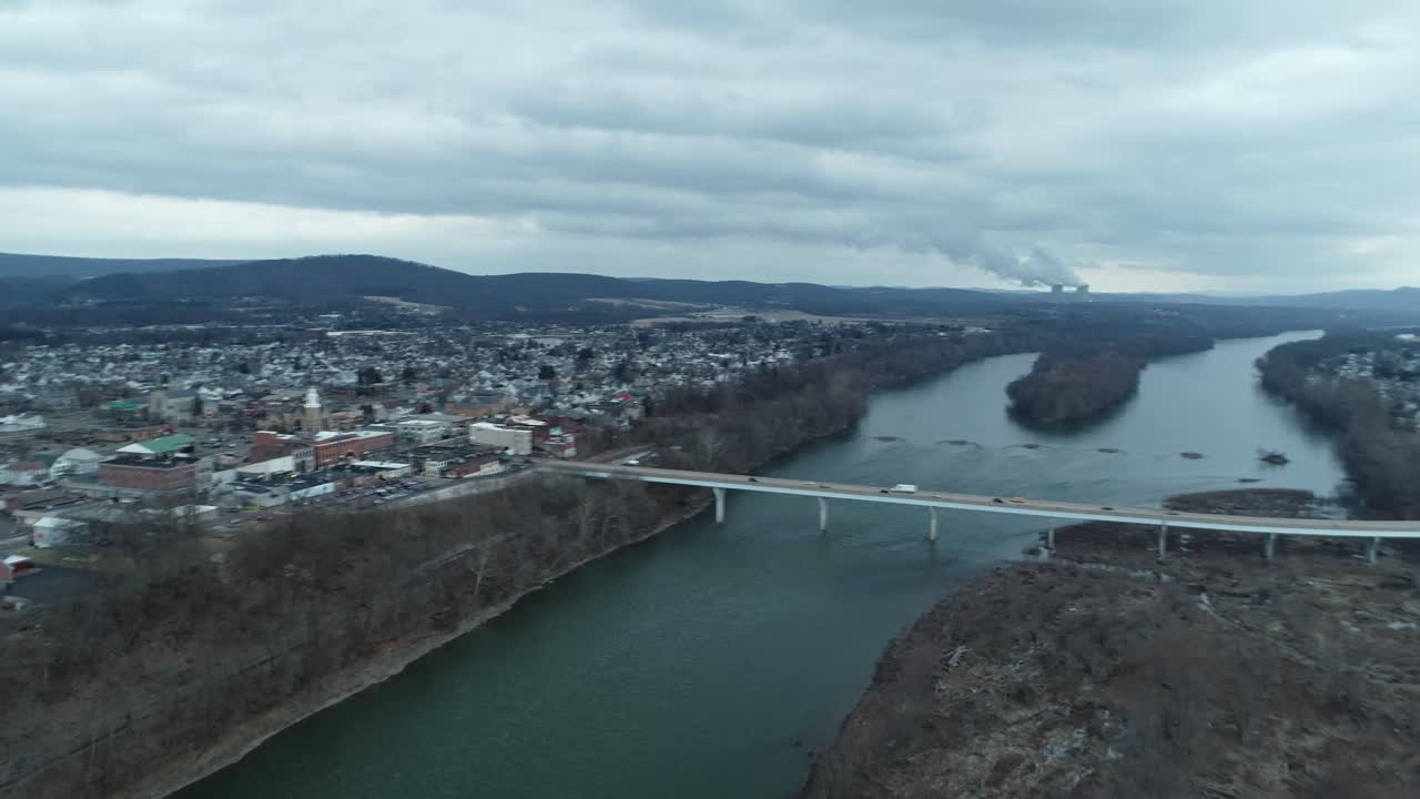 A backward-moving drone shot over a dense city of buildings, houses, cars, and shops, leading toward a scenic lake with a bridge crossing it