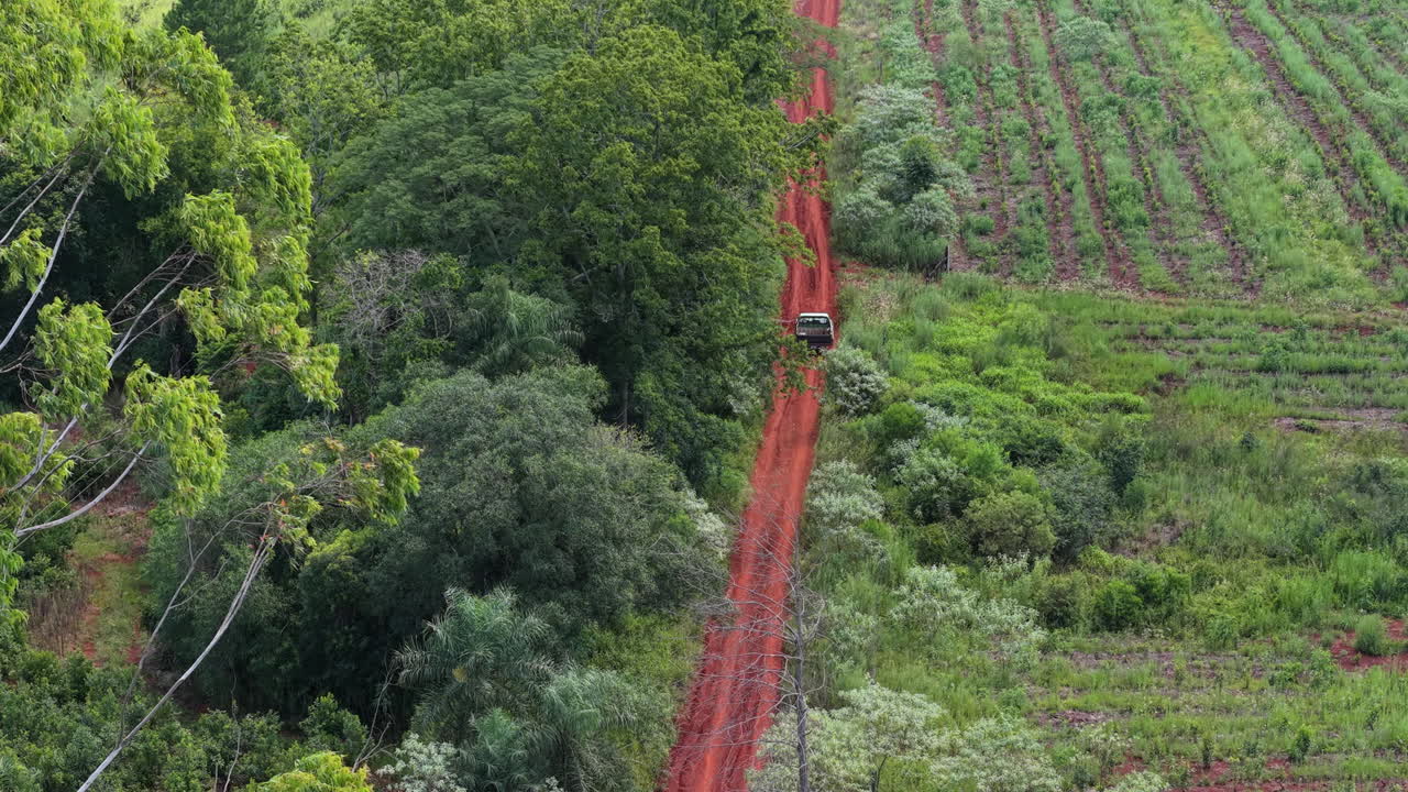 Aerial view of a red dirt road winding through a dense green forest and cultivated fields in Misiones, Argentina, illustrating the intersection of nature and rural life, drone shot