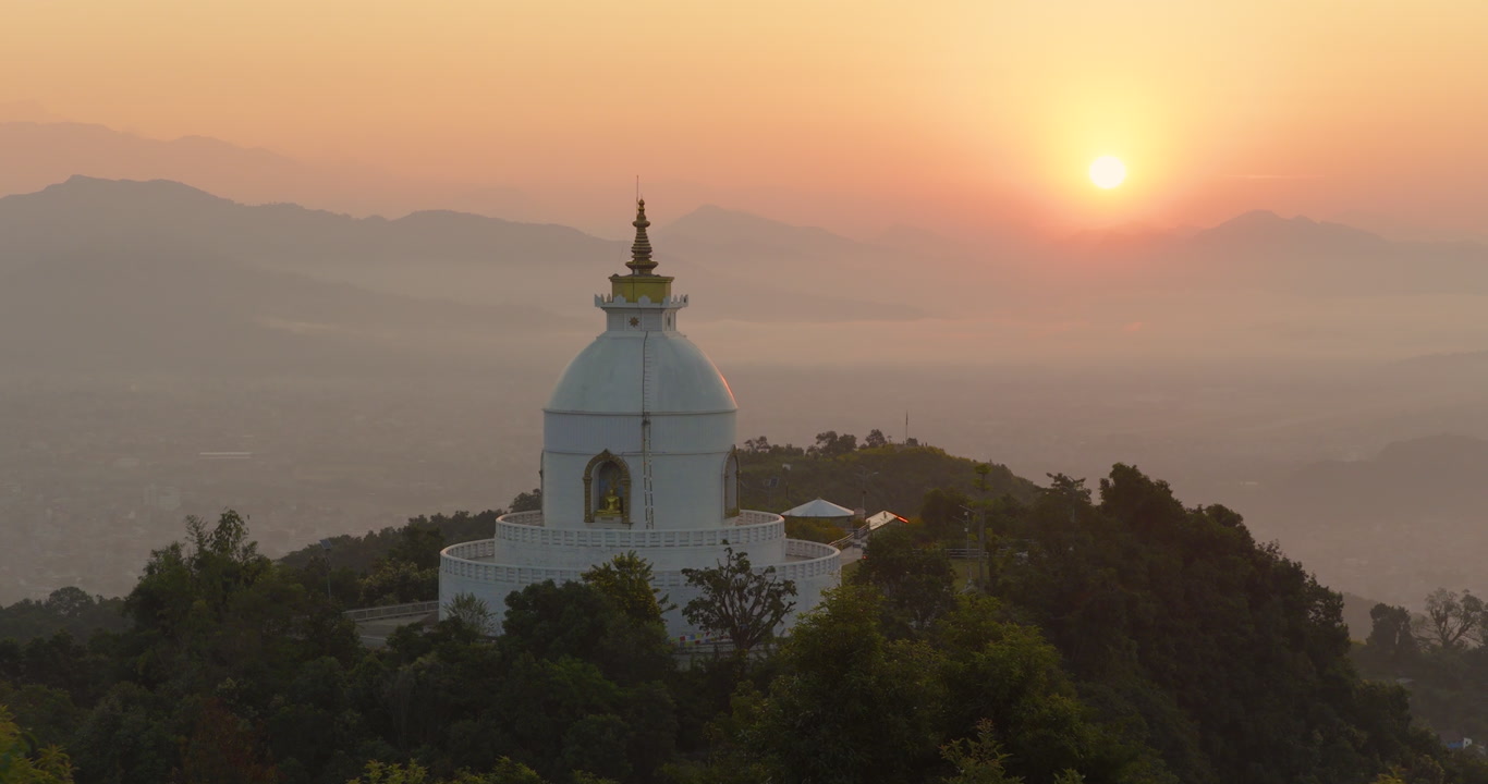 Drone shot of Buddhist monument World Peace Stupa surrounded by lush green forest under golden sky with clouds and warm sunlight in Pokhara Nepal.