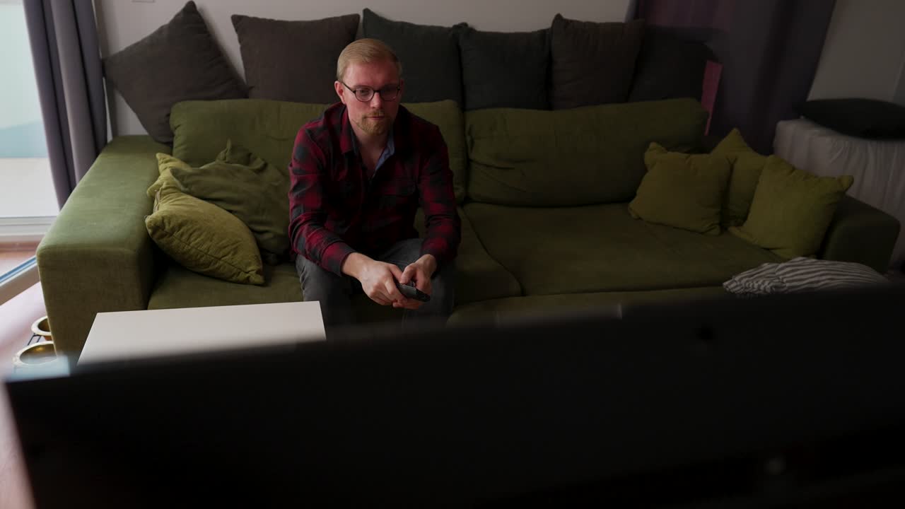 수염을 가진 백인 남자는 소파에 앉아 리모콘을 사용하고 일상적인 집