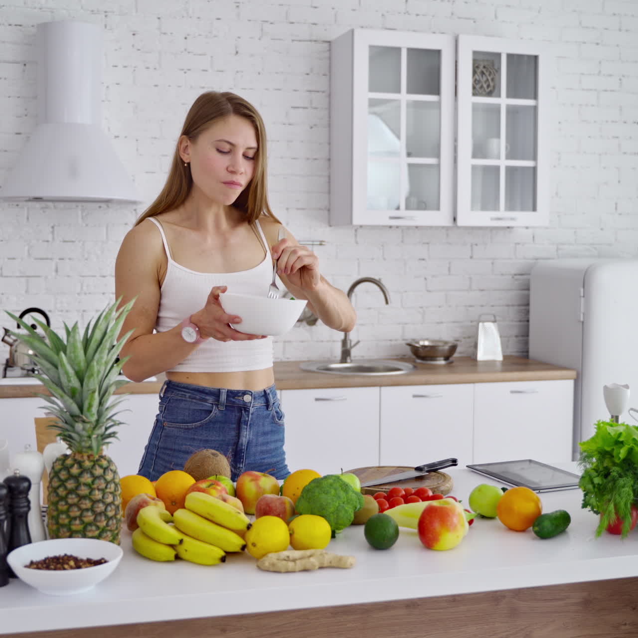 Young woman in the kitchen. Beautiful girl standing near the table with organic food. Lovely female enjoys eating fresh salad on the modern kitchen background. Dieting concept.