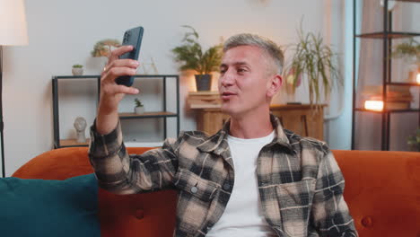 happy young man taking selfie on smartphone communicating video call home online with subscribers