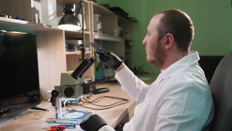 una vista de cerca de un técnico en una bata de laboratorio blanca inspeccionando una placa de circuito, ambos usando el técnico examina la placa de circuito bajo un microscopio, con varias herramientas alrededor