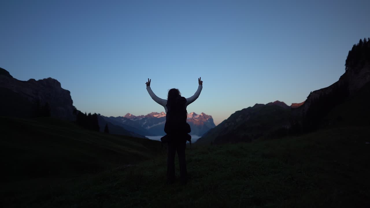 Un escursionista alza le braccia nelle Alpi Svizzere all'alba, esprimendo libertà, realizzazione e solitudine. Scena escursionistica cinematografica sulla Via Alpina, ideale per raccontare storie di avventura, natura e stile di vita all'aria aperta.