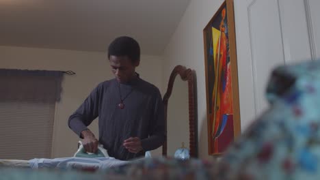 black-man-ironing-shirts-in-bedroom-with-painting-behind-him