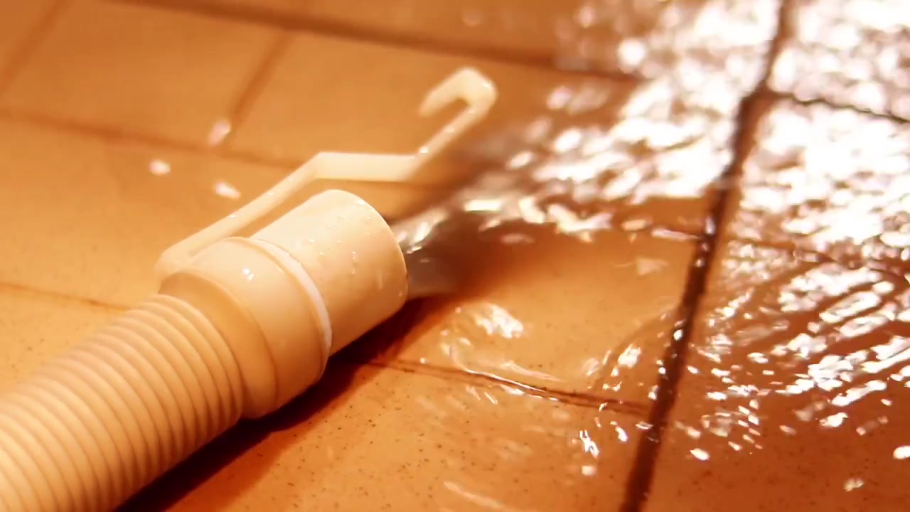 Flexible hose releases a stream of water across a glossy tiled floor in warm indoor light, useful for cleaning, leak, or plumbing concepts.