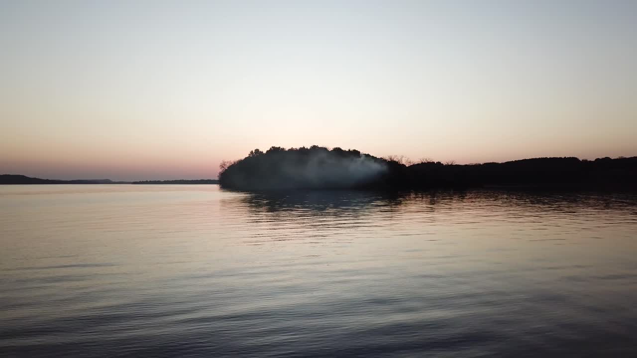 Smoke drifts from a wooded peninsula into the calm waters of Lake Wisconsin under a soft twilight sky. The wide aerial view highlights serene reflections and a mysterious dusk atmosphere.