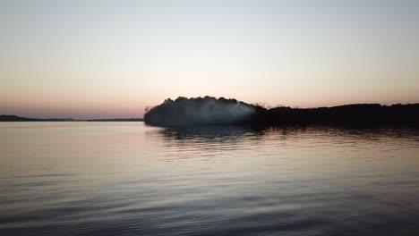 smoke drifts from a wooded peninsula into the calm waters of lake wisconsin under a soft twilight sky. the wide aerial view highlights serene reflections and a mysterious dusk atmosphere.