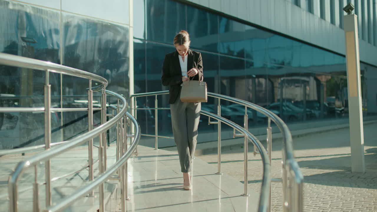 Professional businesswoman leaves work, walking down ramp with metal railings, checking handbag thoughtfully, retrieving keys, modern glass building and parked cars reflecting urban office setting