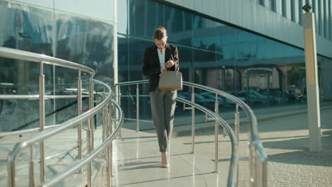 professional businesswoman leaves work, walking down ramp with metal railings, checking handbag thoughtfully, retrieving keys, modern glass building and parked cars reflecting urban office setting