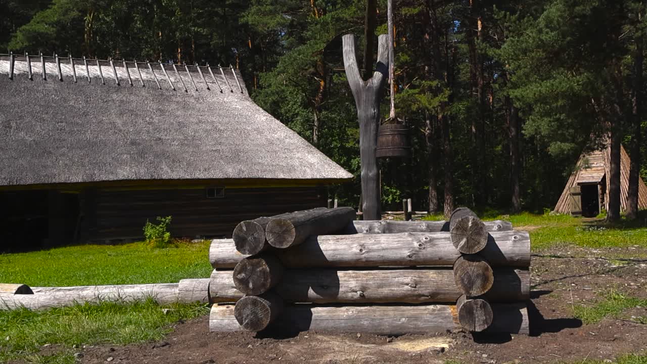 An old historic and vintage wooden well with a wooden bucket hanging above it in a historic farm garden that has log buildings with thached reeds roofing and farn equipment. Pine forest and sunny day