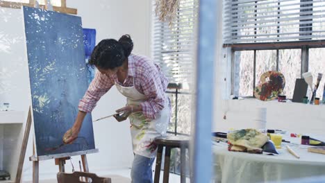 focused biracial female artist in apron painting on canvas in studio, slow motion