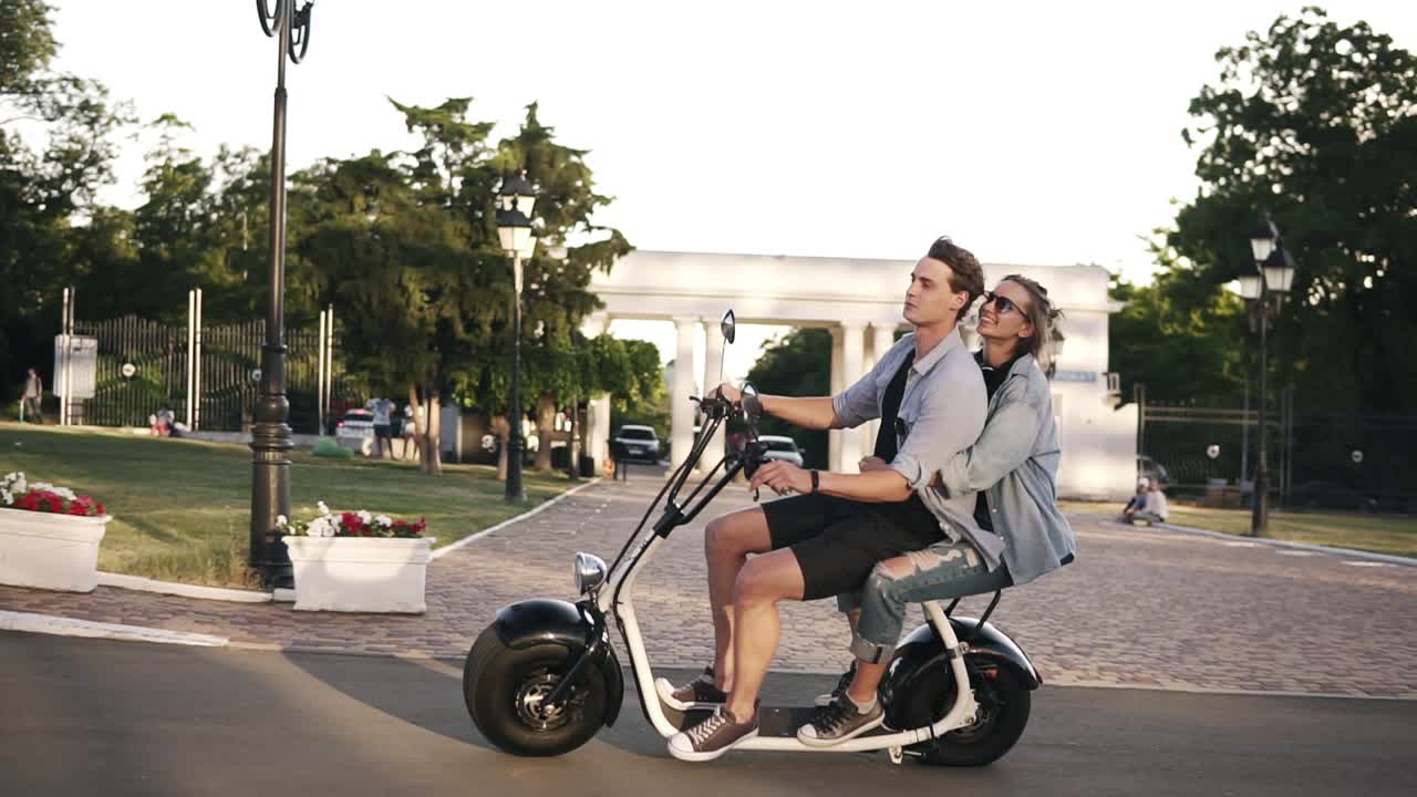 Confident man in blue shirt driving scooter while his girlfriend embracing him. Amazing young woman riding on moped with friend by the parkside