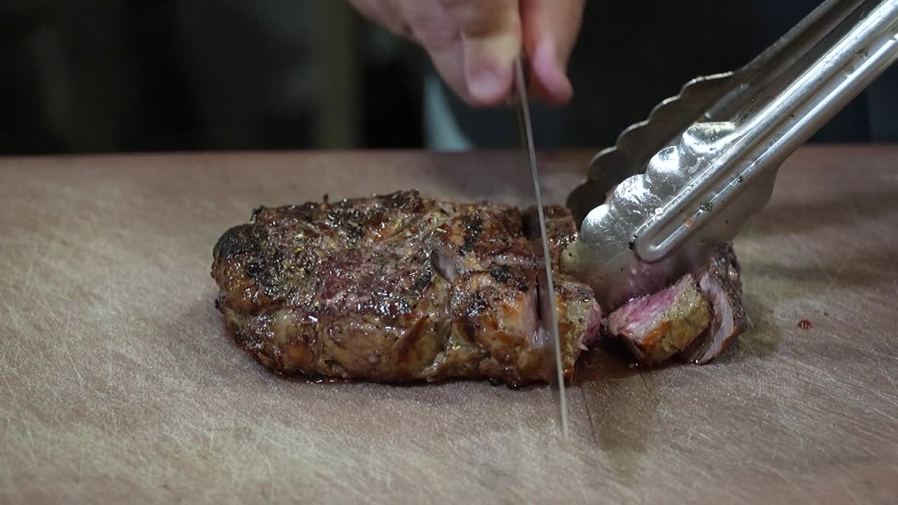 Closeup of Succulent Steak: Knife Cutting on Cutting Board Holding it With Clippers