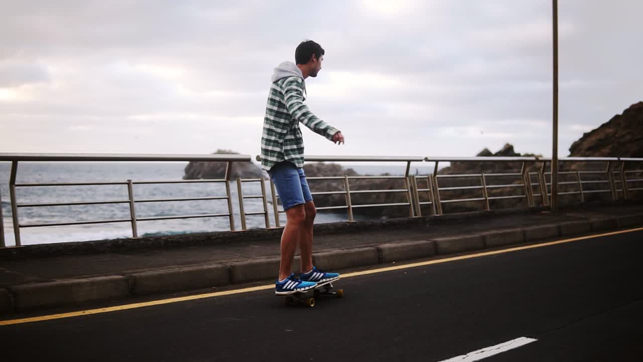 deporte, estilo de vida, extremo y concepto de personas - hombre alto montando longboard por la carretera costera en tiempo nublado. longitud completa de un hombre activo haciendo ejercicio en la carretera. cámara lenta