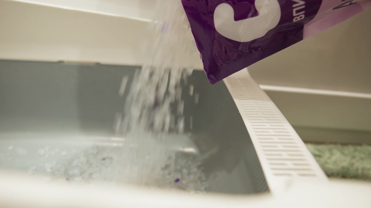 Primo piano, vista dal basso, di detersivo in polvere e in fogli che viene erogato nel cestello aperto di una lavatrice, con una cascata visibile di polvere, frammenti di fogli strappati e particelle galleggianti che creano leggere increspature.