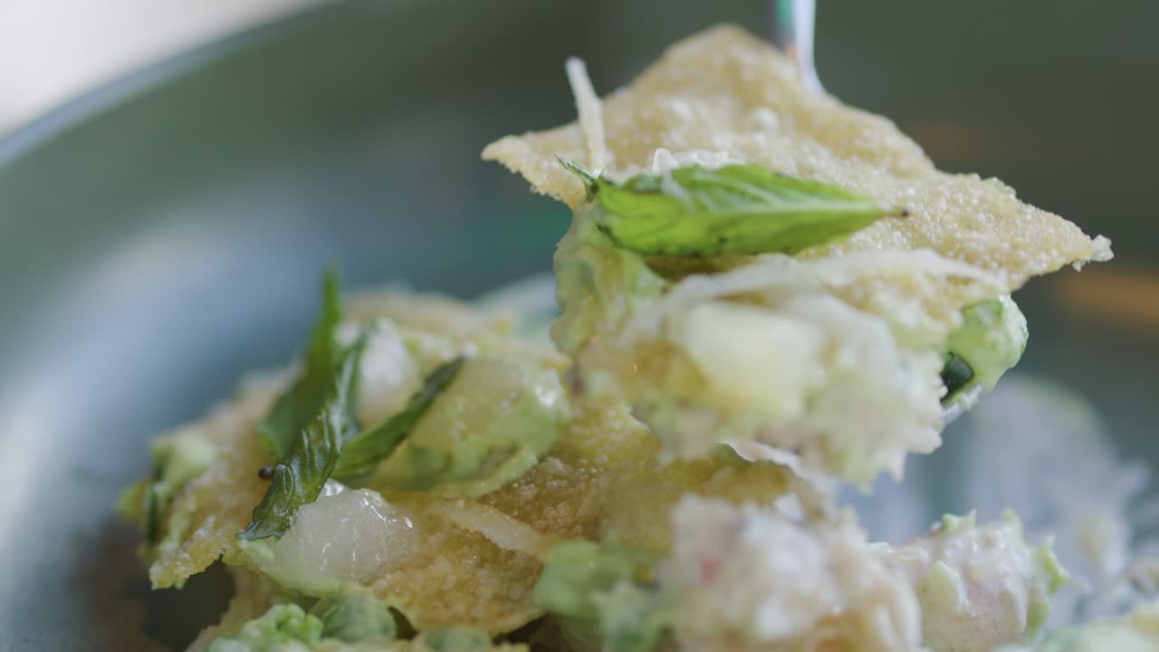 A spoon lifts a portion of crabcake garnished with herbs and avocado from a plate, captured in soft natural lighting with a shallow depth of field