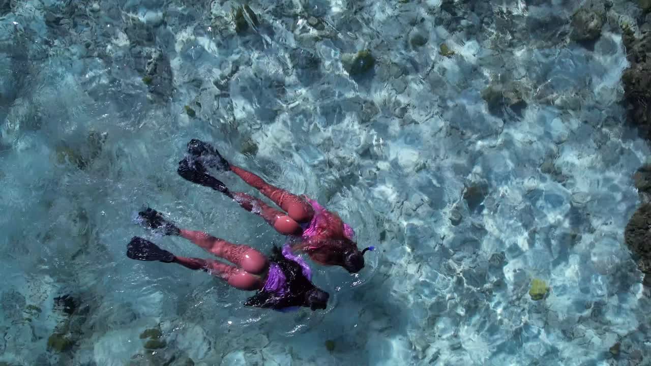 Two women snorkeling over clear blue waters surrounded by coral reefs, top-down aerial view
