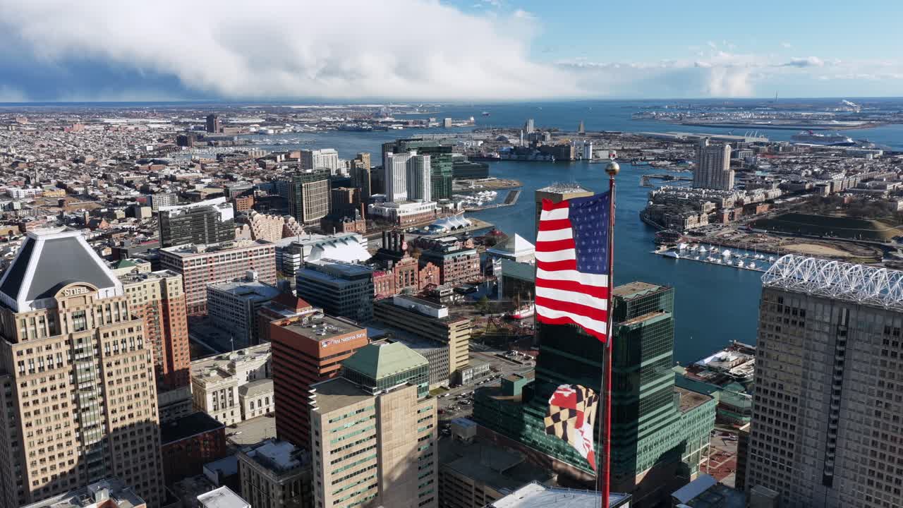 Vista aerea dello skyline del centro di Baltimora con una grande bandiera americana che sventola accanto alla torre della Bank of America, con edifici cittadini, porto e paesaggio urbano sotto nuvole drammatiche.