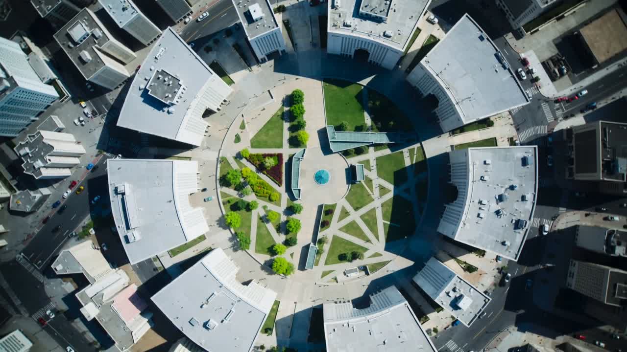 Cámara de dron girando y haciendo zoom sobre una plaza urbana, mostrando una fuente, pasarelas y árboles para planificación.