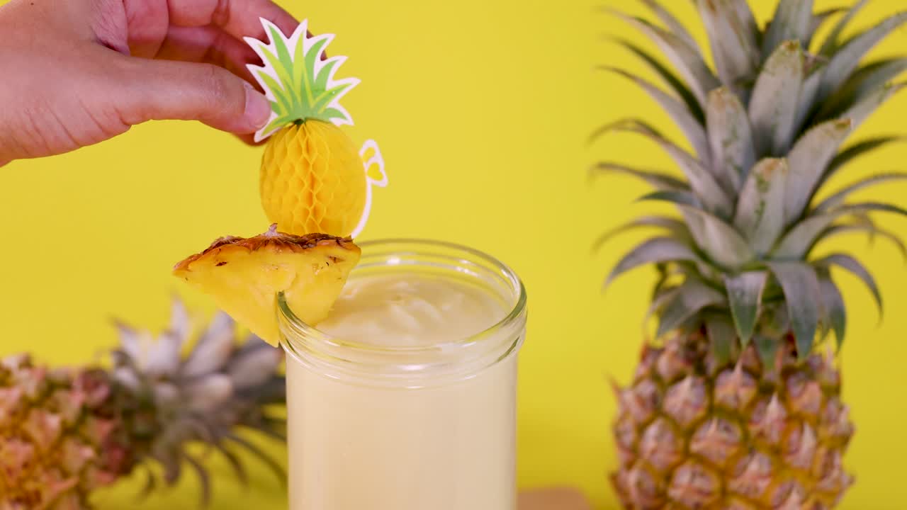 Hand places pineapple skewer garnish on creamy smoothie, bright yellow backdrop, vibrant tropical setting