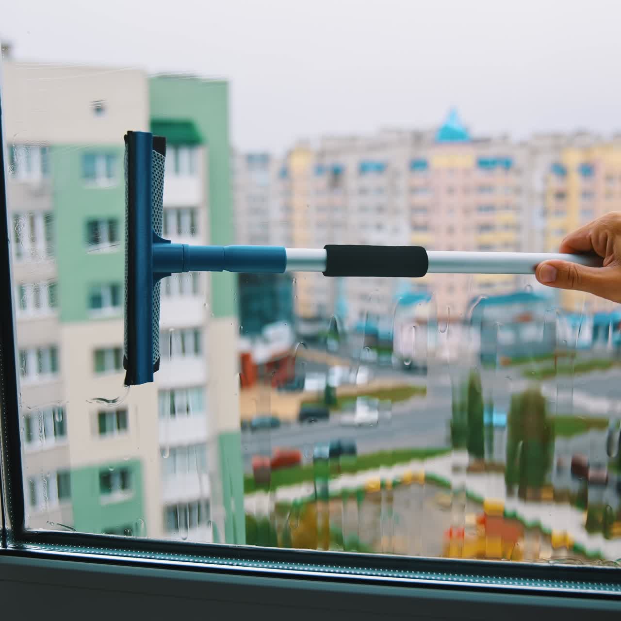 Cleaning window in the city. Man holding household brush and cleans window from outside. View from indoors. Urban buildings background