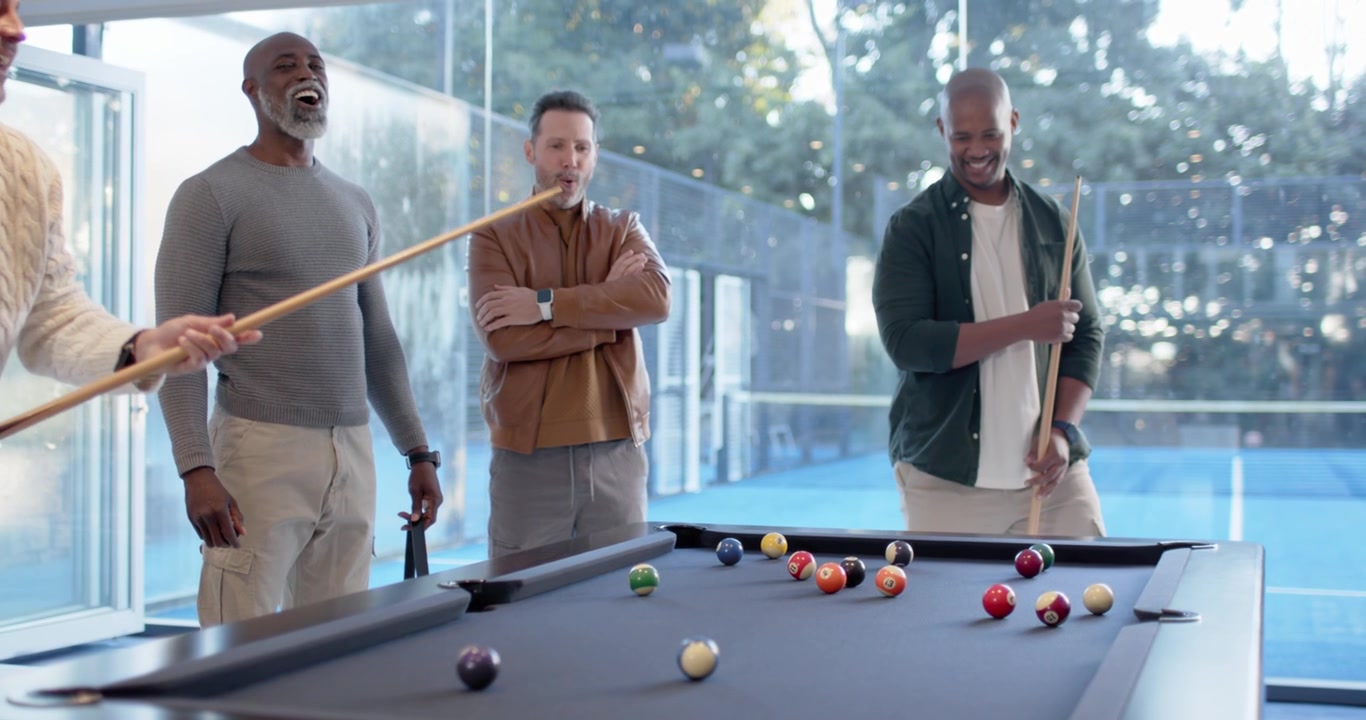 Diverse male friends playing pool at slate-blue table, shooter striking cue ball, balls scattering
