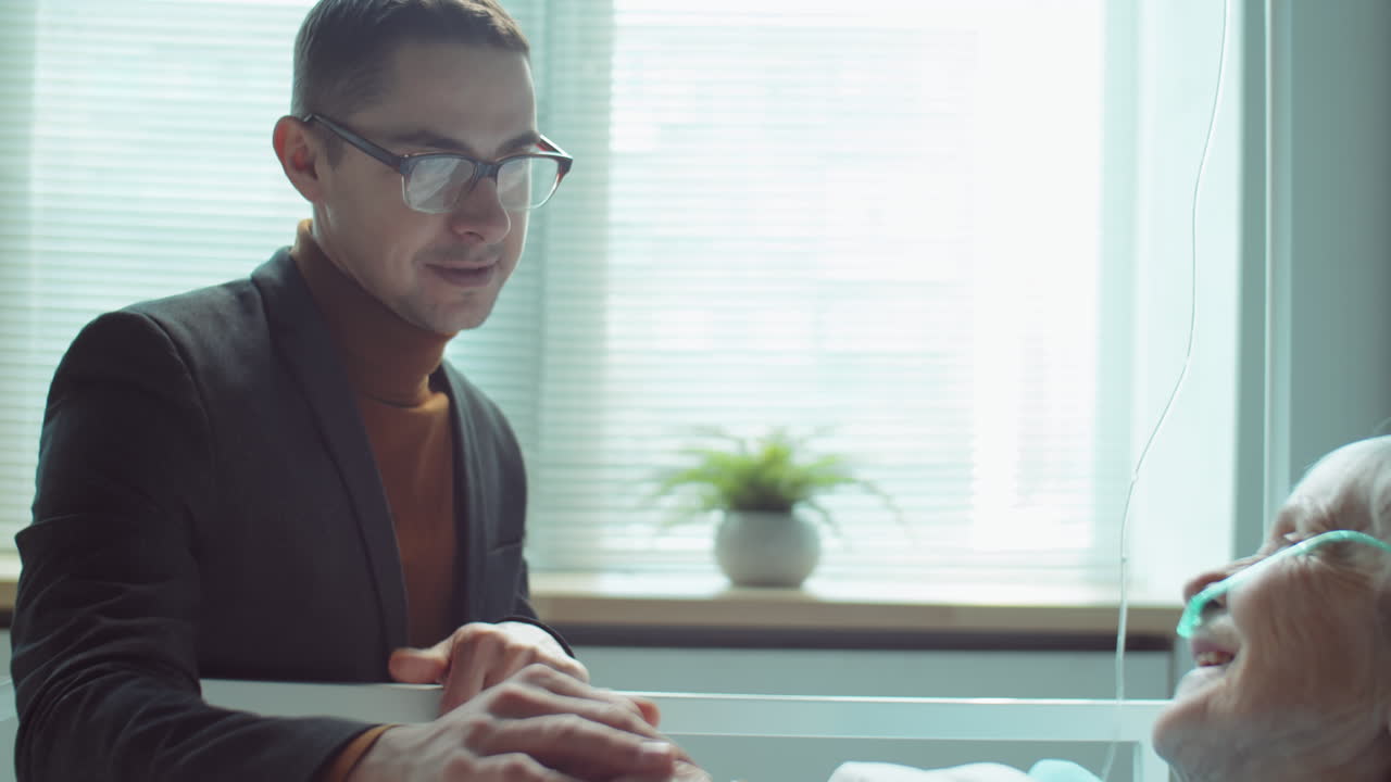 아들이 병원 병실에서 노인 어머니를 지원하고 있습니다.