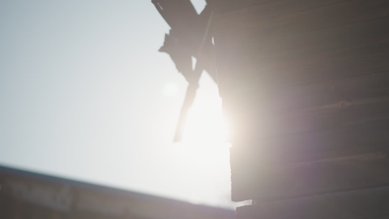 Grondaia di casa illuminata dal sole con sole splendente, silhouette del bordo rotto del tetto in legno, controluce caldo e bagliore morbido, texture rustica e pulviscolo, lenta panoramica a mano che suggerisce la riparazione e l'ispezione di una struttura datata.