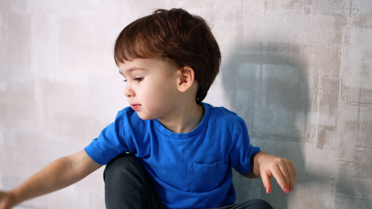 Little kid wearing blue t-shirt sits at the wall. Close up. Lovely toddler plays with a red apple.