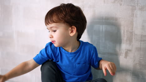 little kid wearing blue t-shirt sits at the wall. close up. lovely toddler plays with a red apple.